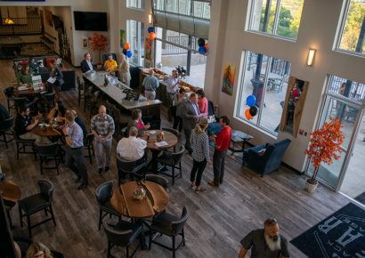 aerial of large event room with cocktail tables and large windows, people gathered in small groups networking at the bar and around the room